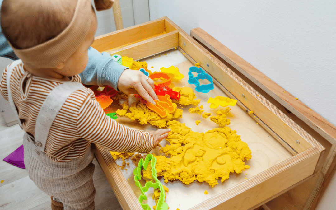 Child playing with yellow dough and plastic cookie cutters in a wooden sensory table.