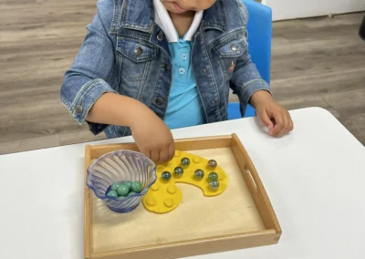 A young girl in a denim jacket plays with a marble board placing marbles on a yellow pegboard inside a wooden tray on a white table